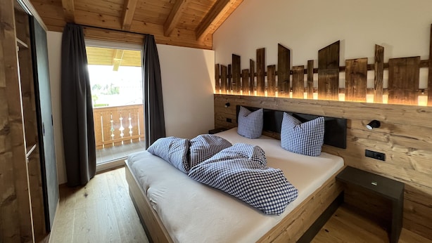 A wooden cabin bedroom with a bed, checkered pillows, a wooden headboard, and a balcony with curtains. - Studlerhof (Oberperfuss)