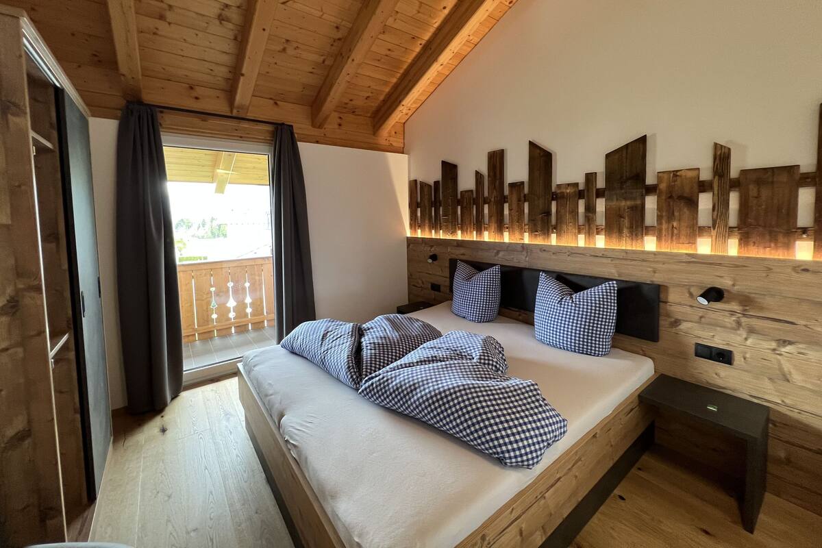 Un dormitorio de cabaña de madera con una cama, almohadillas de cuadros, un cabecero de madera y un balcón con cortinas.