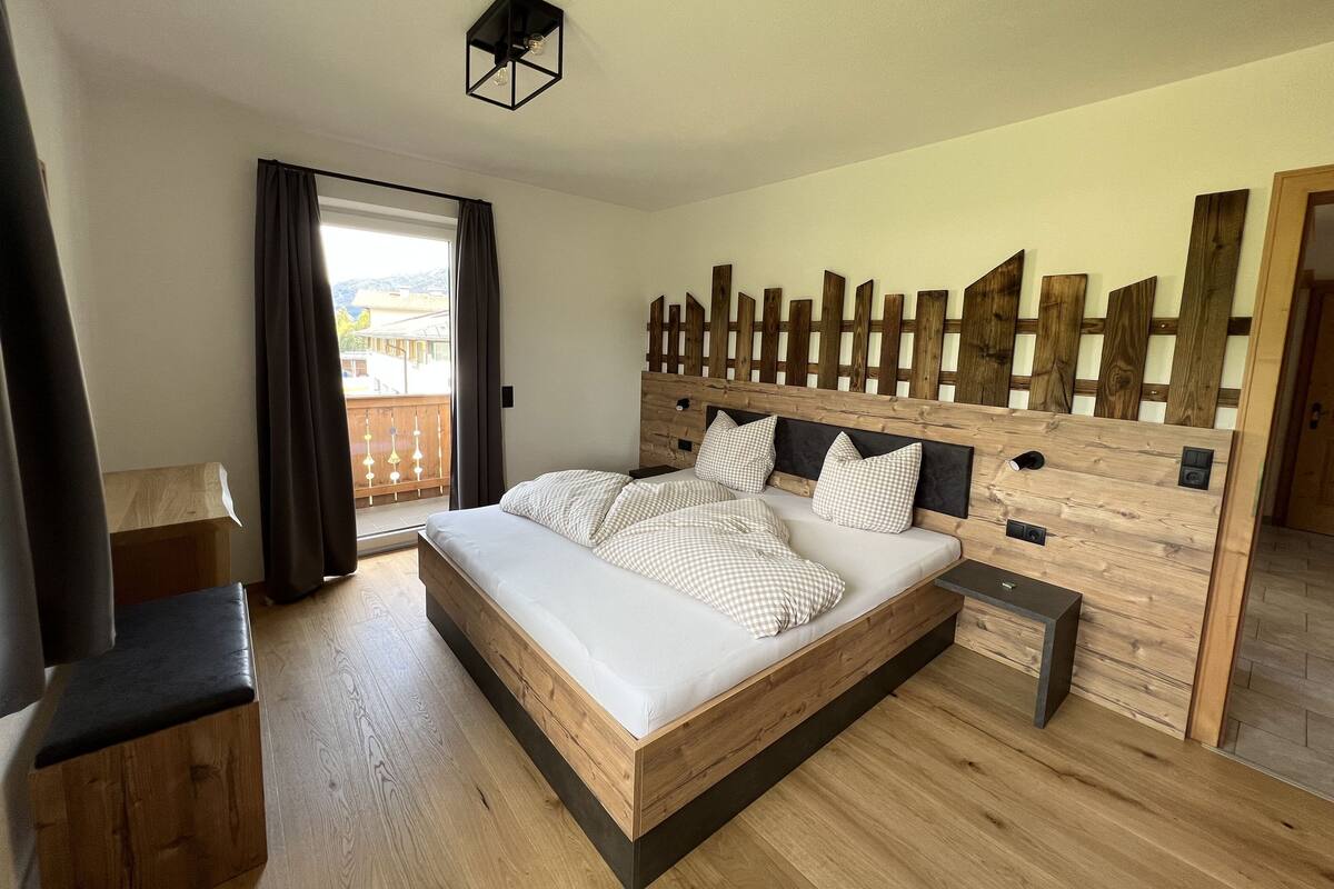 Un dormitorio moderno con un marco de cama de madera, un cabecero con diseño de listones de madera, una mesita de noche y vistas al exterior a través de una ventana con cortinas.