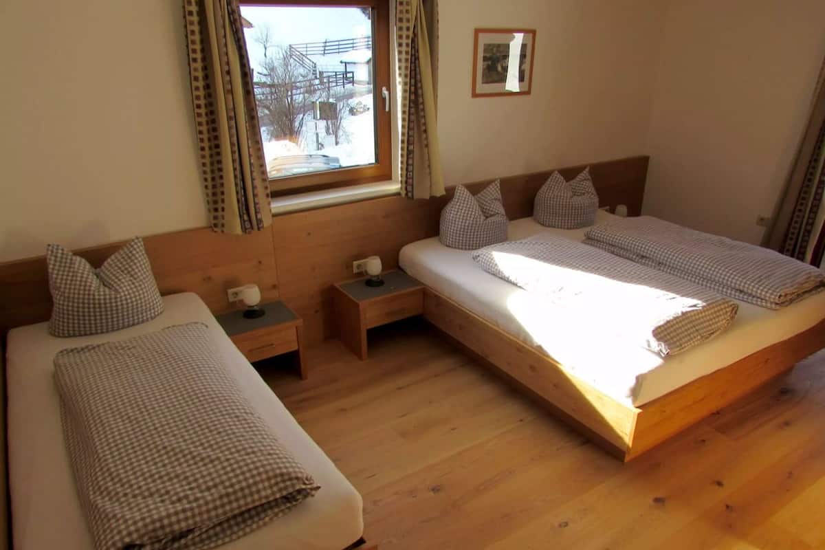 Una habitación con dos camas individuales, cada una con cabecero de madera y almohada a cuadros. En cada cama hay una mesita de noche con una lámpara. Se ve una ventana con cortinas que muestra un paisaje nevado al exterior.