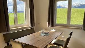 A dining area with a wooden table and four chairs, large windows with curtains, and a view of a green field and mountains. - Studlerhof (Oberperfuss)