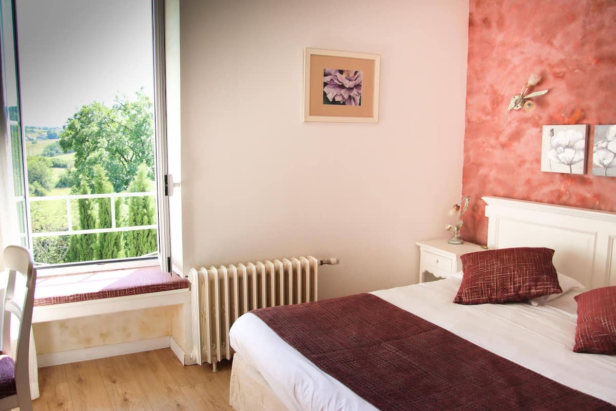 A bedroom with a large window, a radiator, a bedside table, a nightstand, and a bed with pillows.