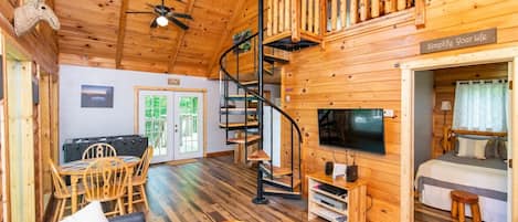 A cozy wooden cabin with a spiral staircase, a dining area, and a bedroom with a bed and TV.
