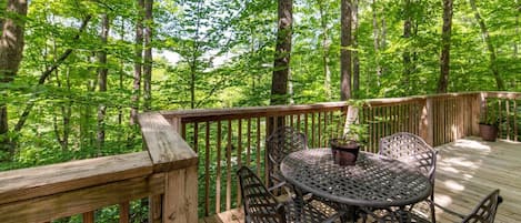 A wooden deck with a round metal table and chairs, surrounded by a forest.