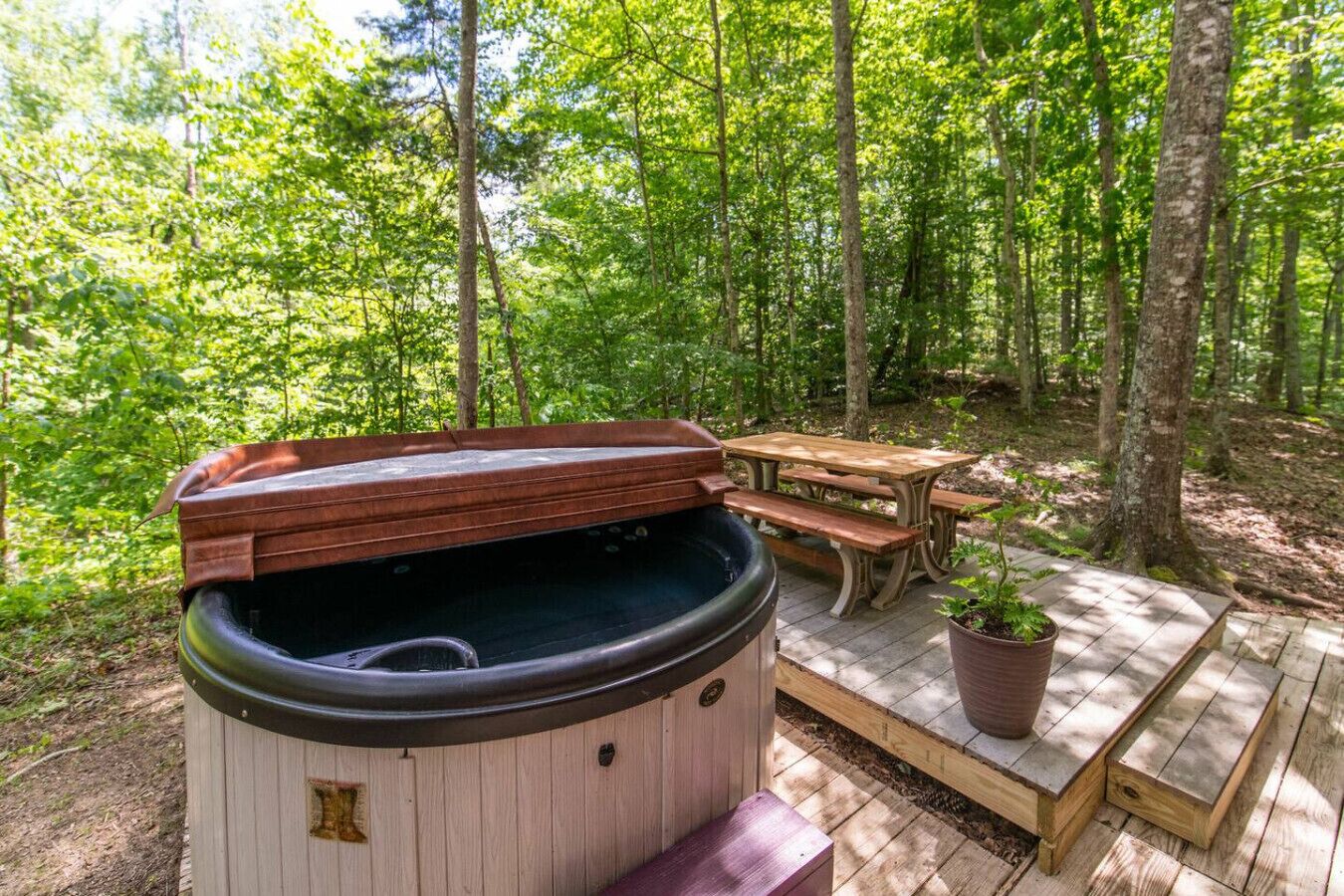 Ett bubbelbad med ett trälock, ett picknickbord och en krukväxt, allt placerat i en skogsmiljö.
