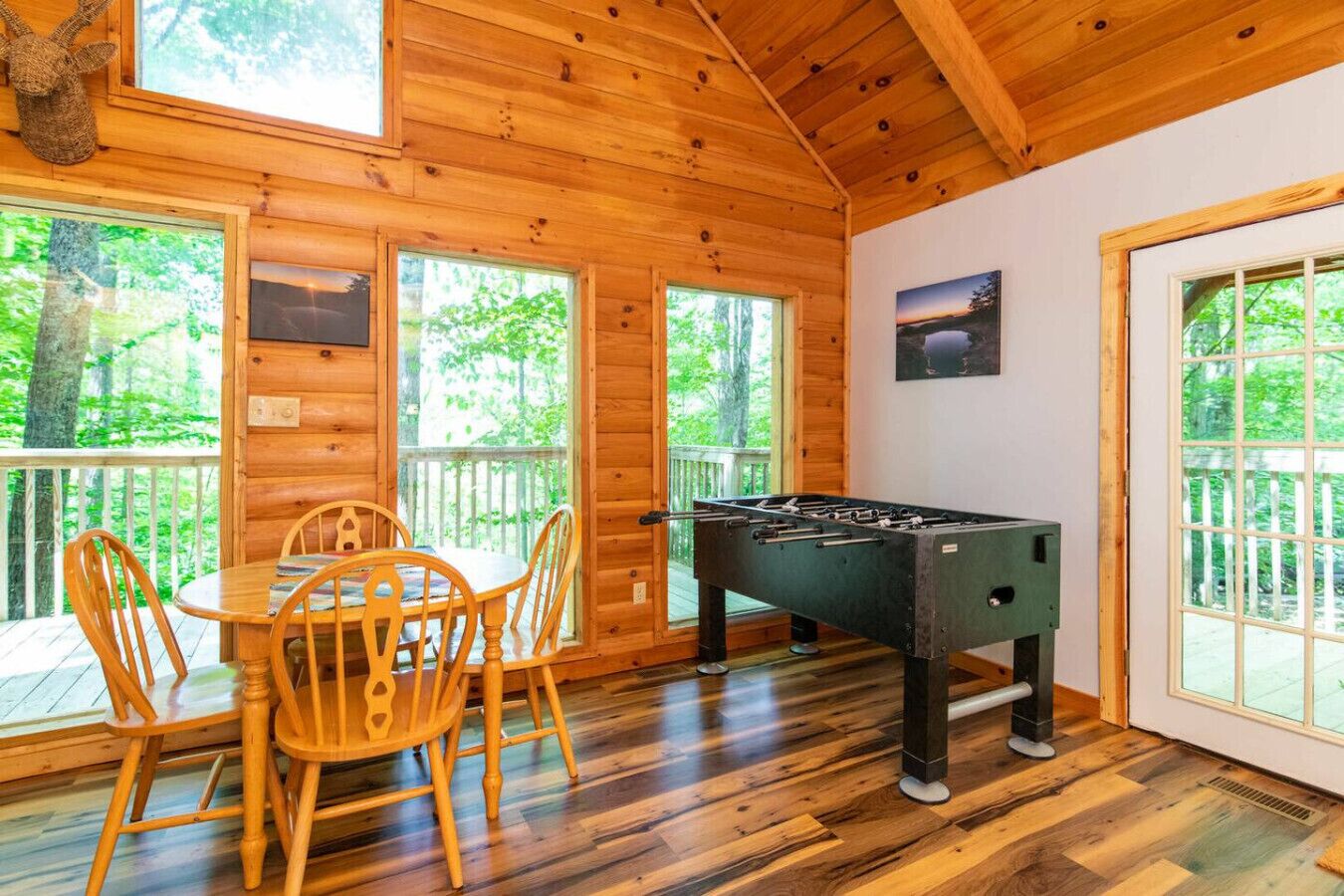 A cozy room with a wooden interior, a foosball table, a dining table with chairs, and large windows offering a view of the outdoors.