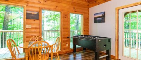 A cozy room with a wooden interior, a foosball table, a dining table with chairs, and large windows offering a view of the outdoors.