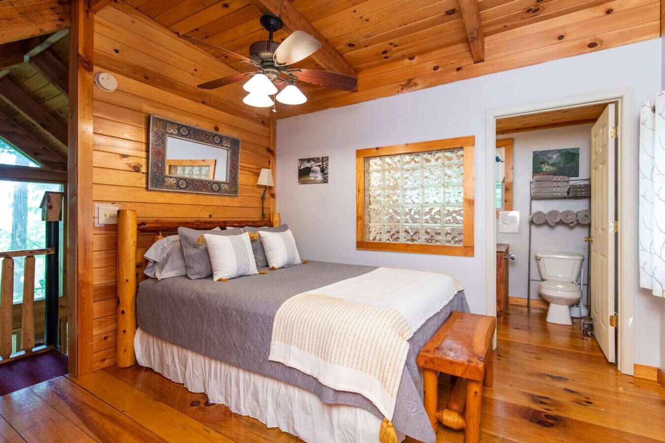 Une chambre de style cabane en bois, avec un lit, un ventilateur de plafond, une fenêtre agrémentée d’un paravent décoratif et une vue sur la salle de bain.
