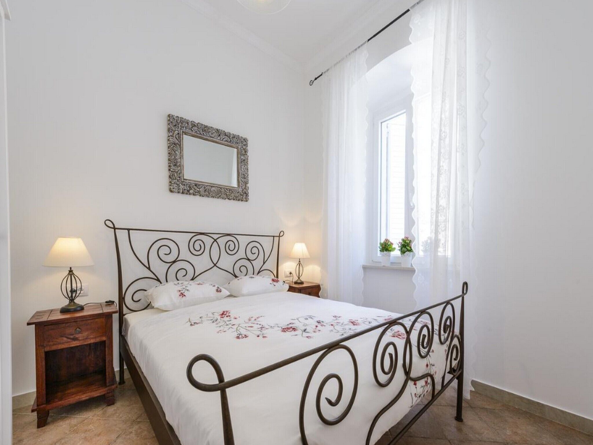 A bedroom with a bed, two bedside tables with lamps, a window with curtains, and a mirror on the wall.