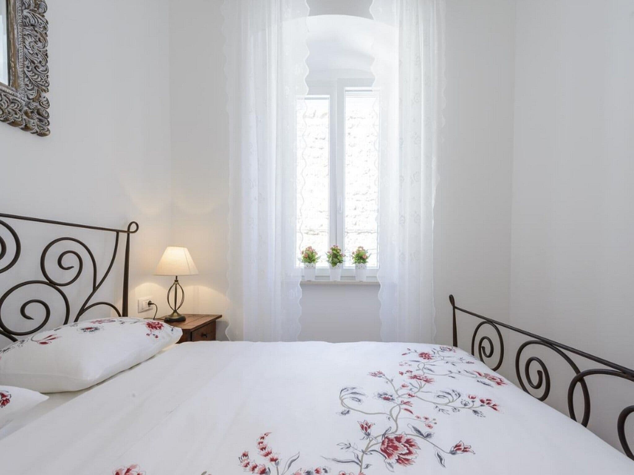 A bedroom with a bed, a bedside table with a lamp, a window with curtains, and a wall-mounted mirror.