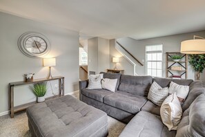Een woonkamer met een hoekbank, een ronde wandklok en een trap naar een hoger niveau.