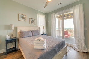 A bedroom with a bed, a nightstand, a lamp, a window with curtains, and a view of the outside. - Beachfront Oasis w/ 2 Large Decks, BBQ & Views! (Topsail Beach)