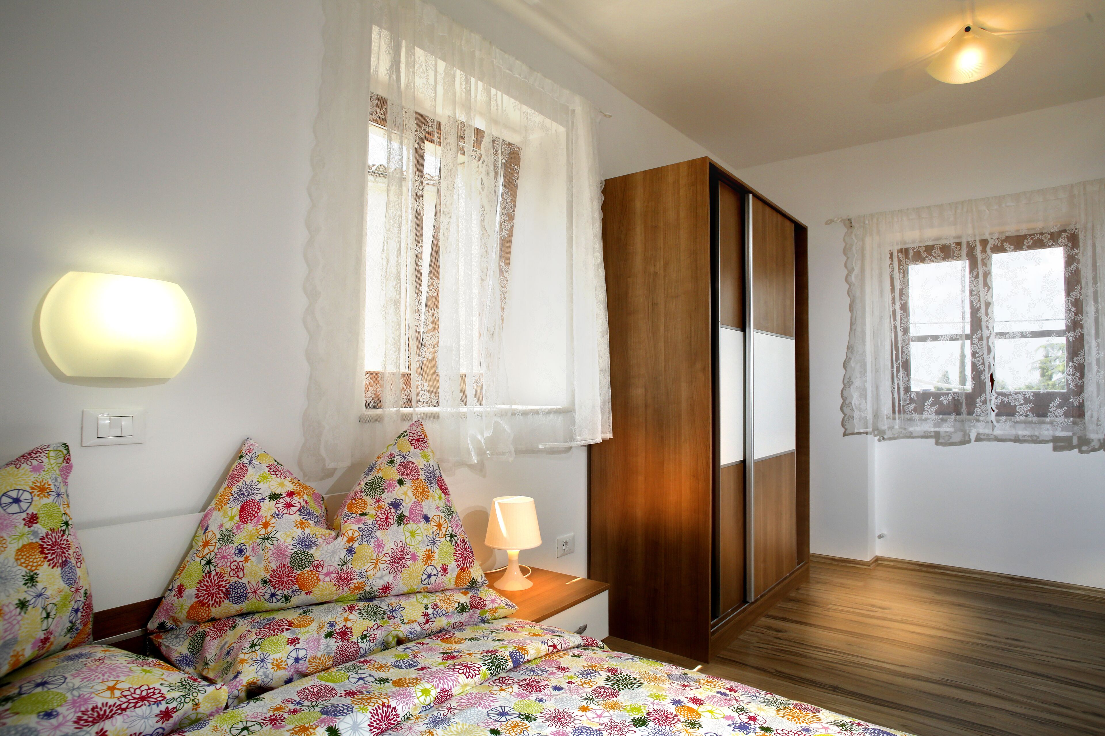 Une chambre  à coucher comprenant un lit, une table de chevet, une lampe, une armoire et deux fenêtres avec des rideaux.