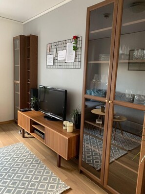 A living room with a TV on a wooden stand, a glass partition, a wall-mounted bulletin board with papers, and a patterned rug. - StayPlus Central 1BR  Apartment (Kristiansand)