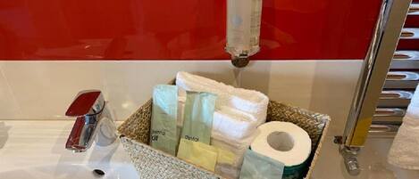 A bathroom sink with a bottle of hand soap, a toothbrush, and a basket containing a towel, a toothbrush, and a roll of toilet paper.