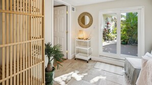 A room with a wooden partition, a white door, a round mirror, a white shelf with a lamp, and a view of a garden through a large window.