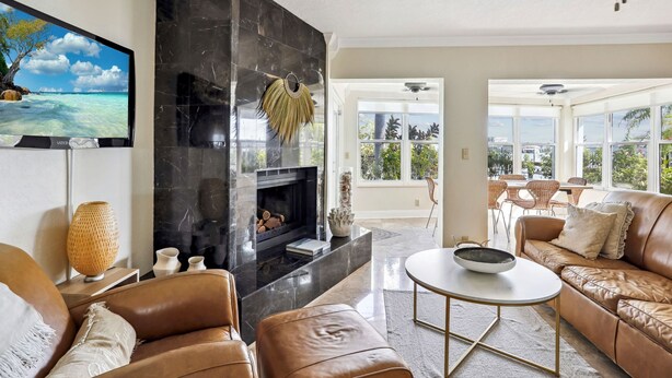 A modern living room with a fireplace, leather sofas, a flat-screen TV, and large windows offering a view of the outdoors.