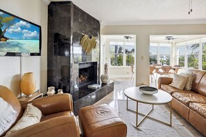 Ein modernes Wohnzimmer mit Kamin, Ledersofas, einem Flachbildfernseher und großen Fenstern mit Blick ins Freie.