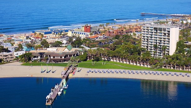 Ein Hotel an der Küste mit mehreren Etagen, ein Strand mit Sonnenschirmen und einem Steg, der ins Wasser führt.