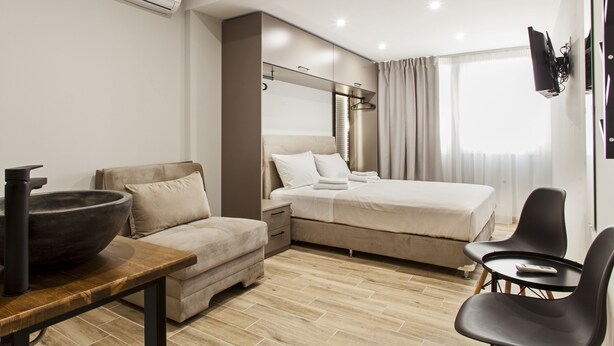 A modern hotel room with a bed, a sofa, a table with a bowl, and a TV mounted on the wall. - Raise The Heraklion Project (Heraklion)