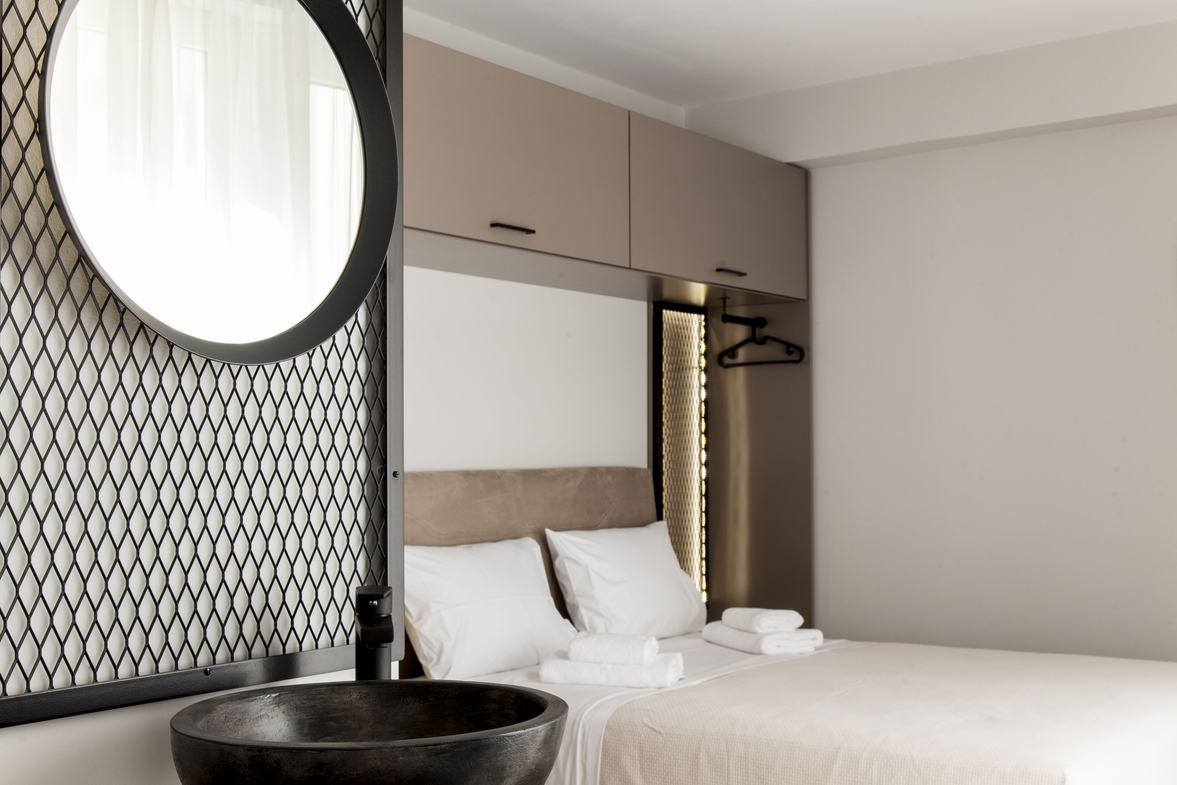 Ein modernes Hotelzimmer mit einem großen, runden Spiegel, einer Holztasse, einem Bett mit weißen Bettwäsche und einem Schrank mit Kleiderstange.