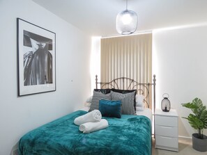 A neatly made bed with a teal bedspread and white towels, a metal-framed headboard, a bedside table with a lantern, and a framed picture on the wall.