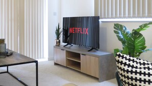 A living room with a TV on a stand, a sofa, a coffee table, and a potted plant. - Tudors eSuites Apartment with 4 Beds (Birmingham)
