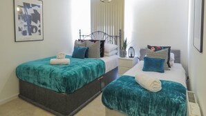 A bedroom with two beds, each with teal bedding and multiple pillows. There is a framed abstract painting on the wall, a bedside table with a lamp, and a window with blinds. - Tudors eSuites Apartment with 4 Beds (Birmingham)