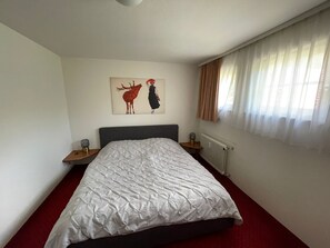 A bedroom with a bed, a nightstand, a wall art piece, a window with curtains, and a radiator. - Ferienwohnung Schwarzwald (Hinterzarten)