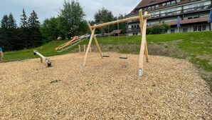 Un terrain de jeux avec des balançoires et un toboggan, entouré d’une zone recouverte de gravier, avec un bâtiment visible au loin.