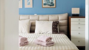 A neatly made bed with a beige headboard, a white quilt, and pink towels. There are two framed pictures on the wall above the bed.