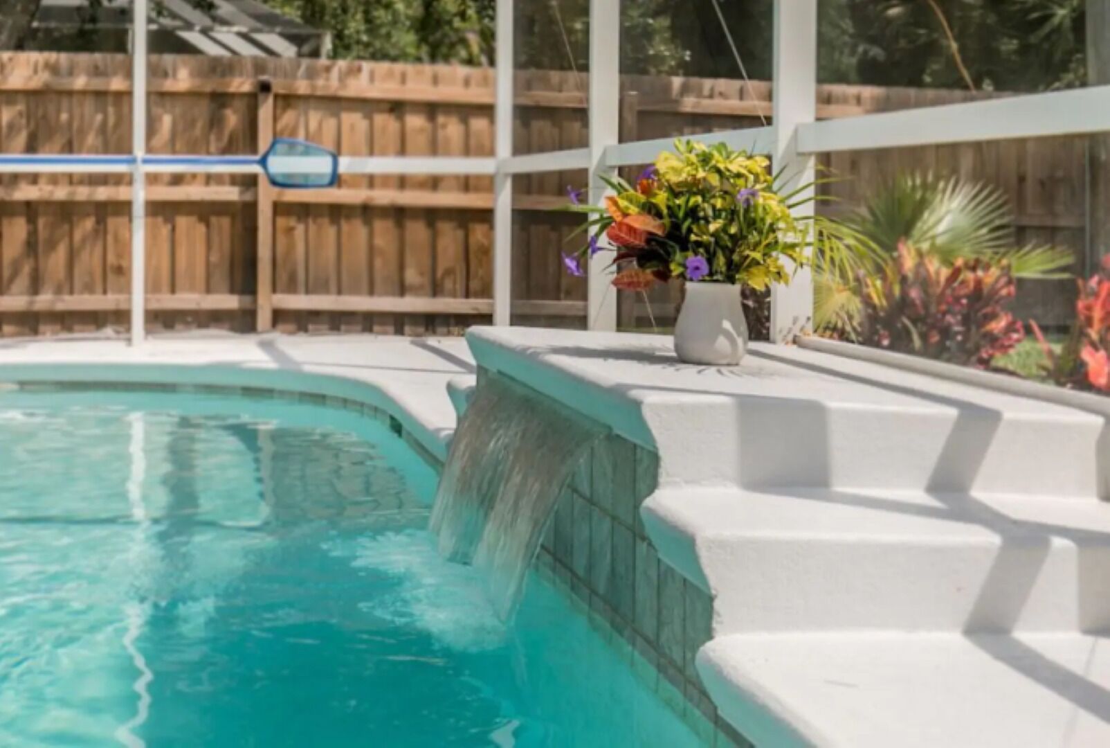 A swimming pool with a waterfall feature, surrounded by a wooden fence and a white deck.