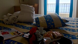 A hotel room with a bed, towels, a magazine, a tray with food and drinks, and a view of the sea through a window.