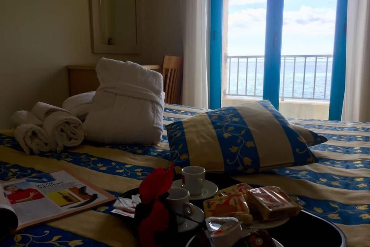 A hotel room with a bed, towels, a magazine, a tray with food and drinks, and a view of the sea through a window.