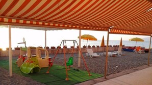 A beachside play area with a swing set, slides, and beach chairs under striped canopies.