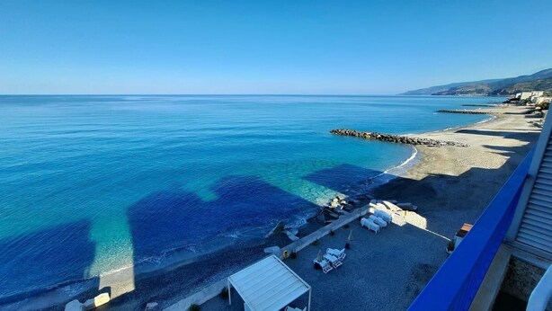 A beach with clear blue water, a rocky breakwater, and a row of white lounge chairs along the shore.