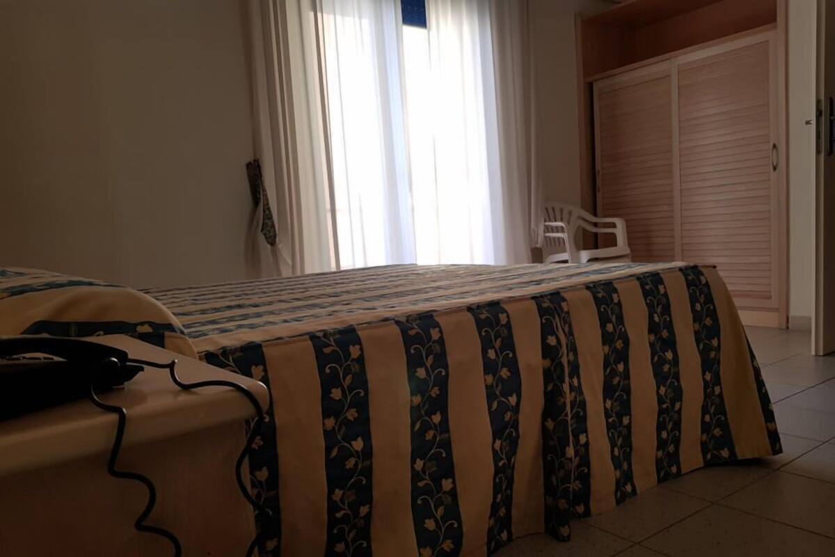 A bedroom with a bed, a bedside table with a telephone, a window with curtains, and a wardrobe.