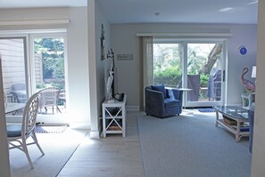 A spacious living room with a large sliding glass door leading to a garden, a blue armchair, and a white side table with decorative items.