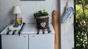 Una habitación con un cuadro enmarcado de frutas y flores en la pared, una mesita con una lámpara, una cesta con etiqueta en forma de corazón y un bolso colgado.