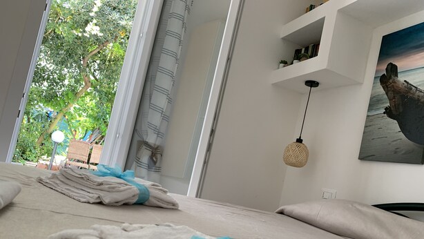 Una cama bien hecha con toallas y un lazo azul, una lámpara colgante de fibra natural y una estantería con libros y adornos.