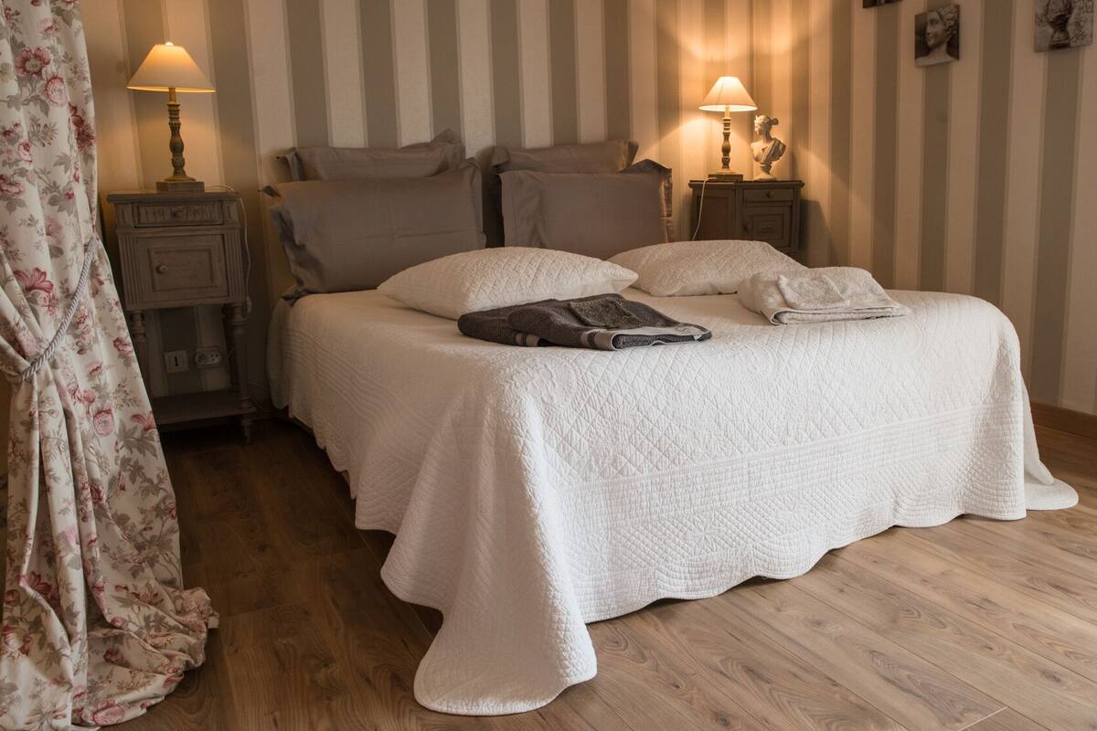 Una cama bien hecha con un edredón blanco acolchado, dos lámparas de noche y una cortina estampada con motivos florales.