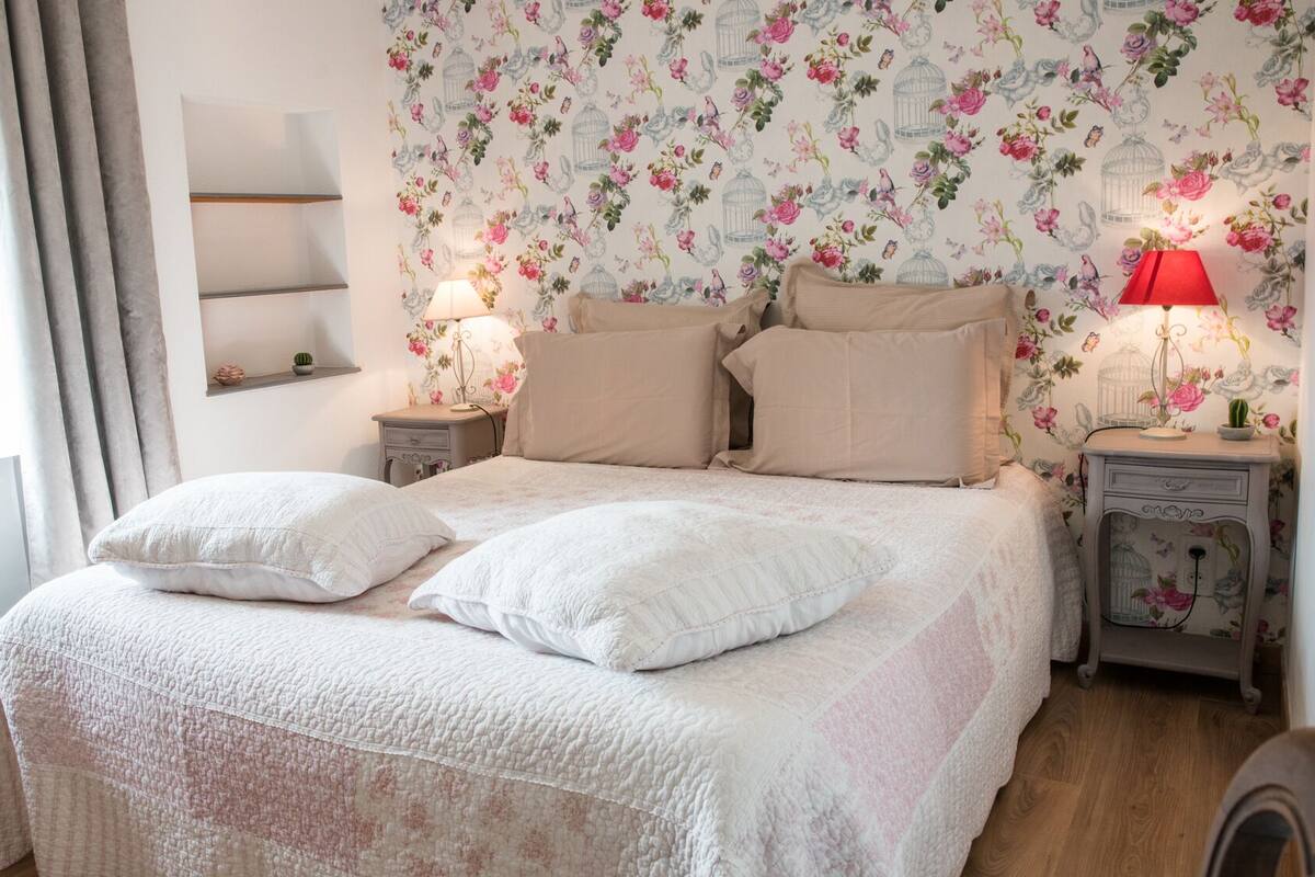 Un dormitorio con una cama grande, dos mesitas de noche, una mesita auxiliar, un ventanal con cortinas y un papel tapiz floral.