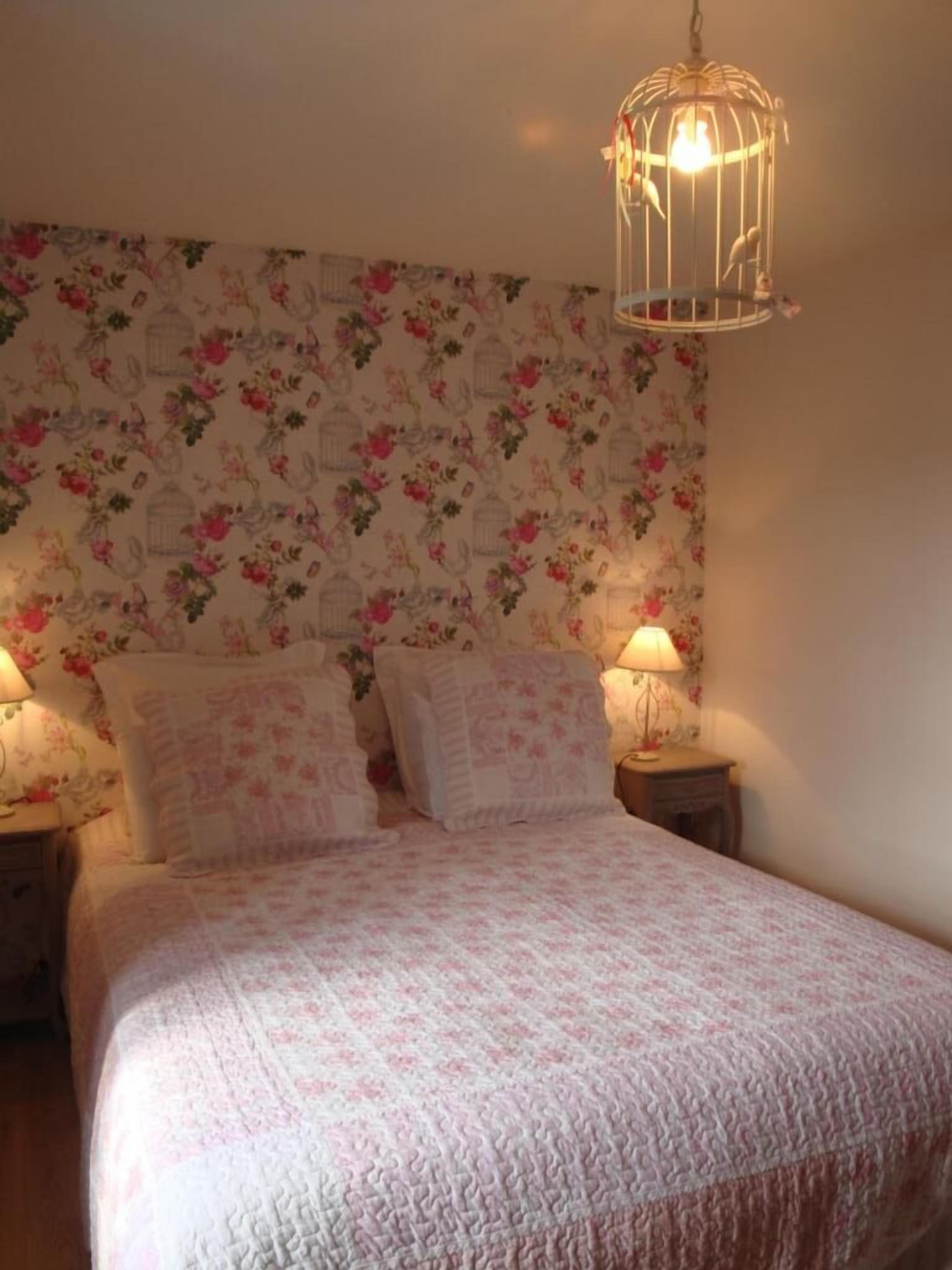 Une chambre à coucher avec un papier peint à motifs floraux, un lit recouvert d’une couvre-lit à motifs, deux lampes de chevet et un luminaire suspendu.