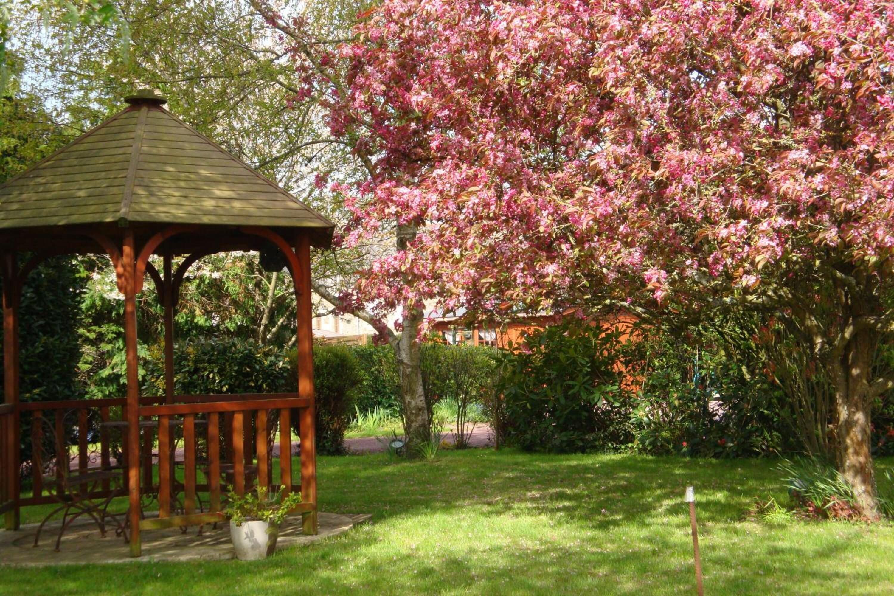 A gazebo in a garden with blooming pink flowers and green grass.