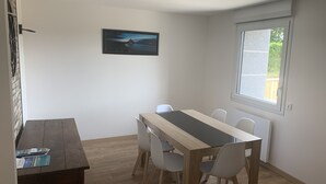 Een eethoek met een houten tafel en witte stoelen, een lijst met een foto aan de muur, en een raam met uitzicht op groen.
