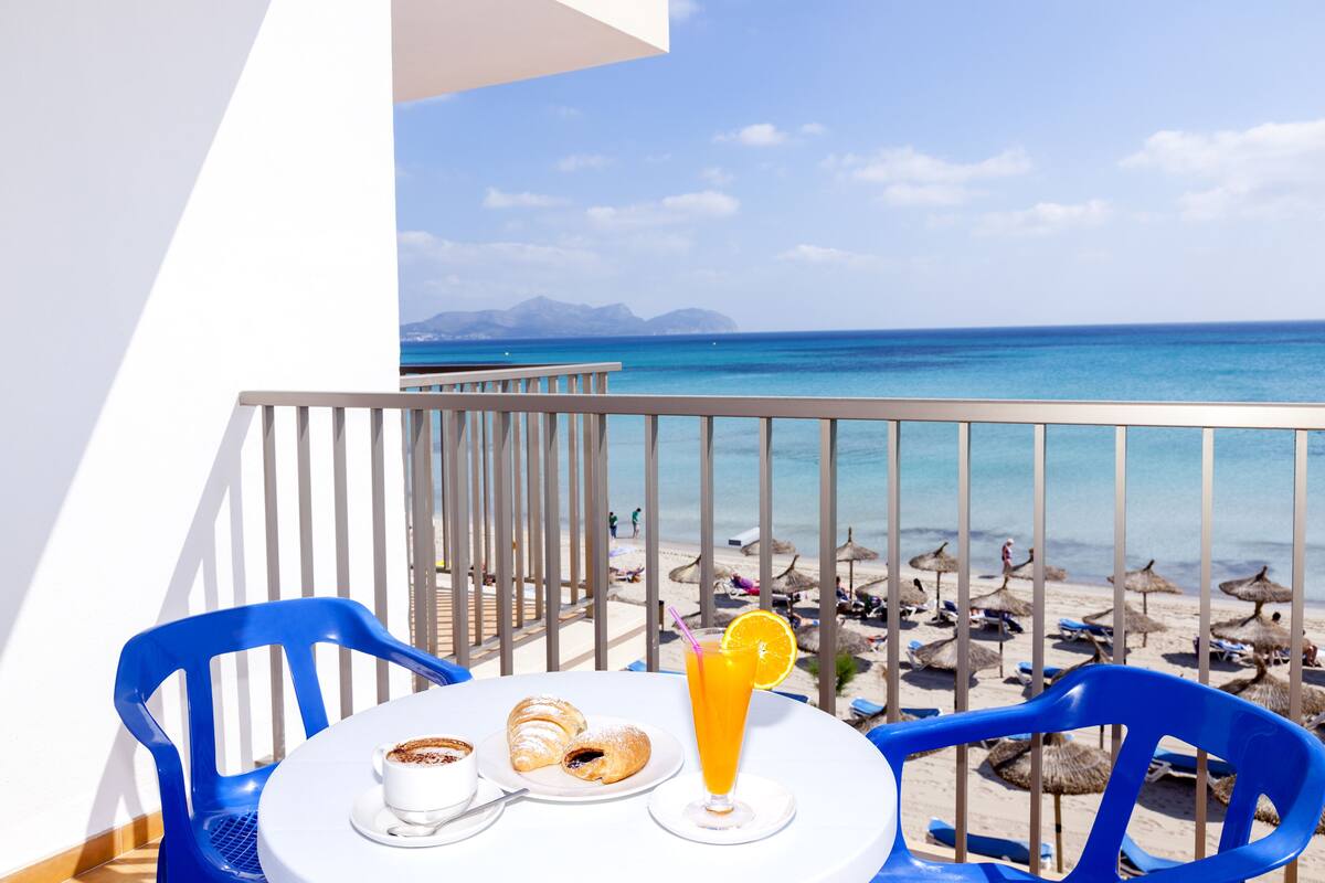 Un balcón con una mesa dispuesta para el desayuno, con vista a una playa con sombrillas y gente.
