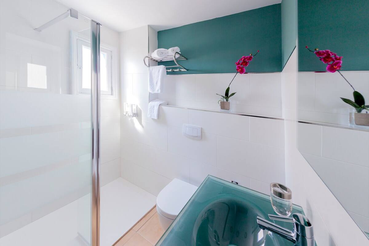 Un baño moderno con cabina de ducha de vidrio, azulejos blancos, una pared de acento en color turquesa, un lavamanos de vidrio y dos orquídeas en macetas.