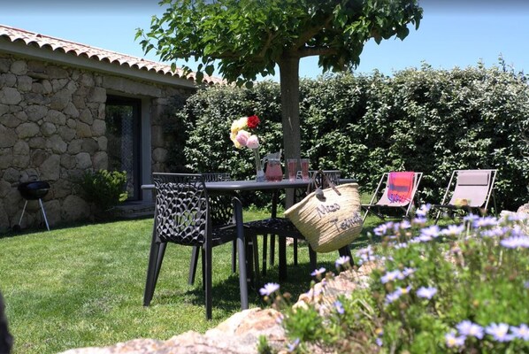 A patio with a stone wall, a table with chairs, a grill, and a tree providing shade. - Rosumarinu Apartment (Figari)