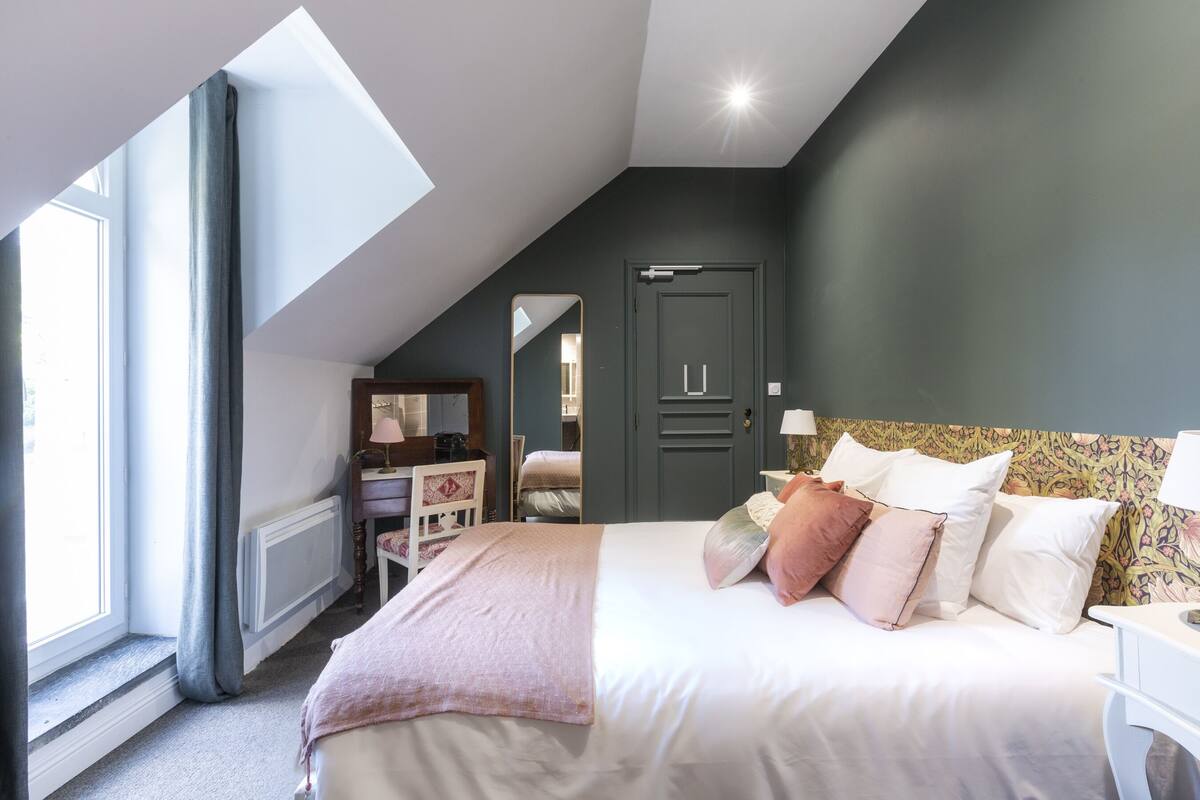 A bedroom with a sloped ceiling, a bed with white and pink bedding, a dresser, a mirror, and a door with a 'U' symbol.