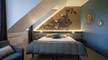 Une chambre avec un plafond en pente, un lit recouvert d’une couette grise, une table de chevet, une chaise et un miroir fixé au mur.